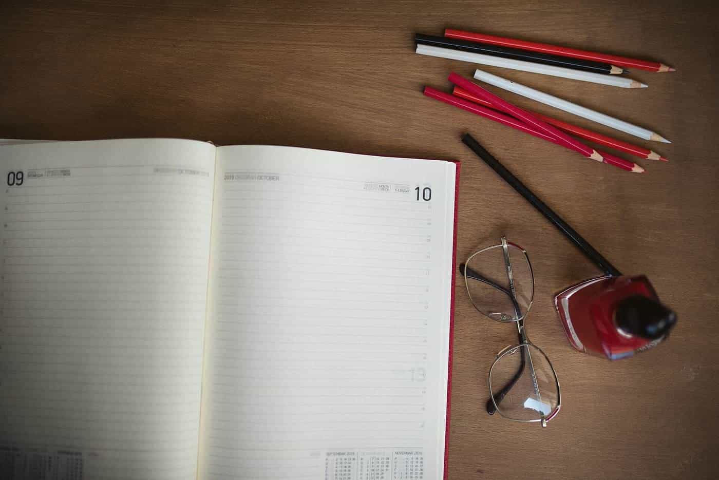 An open diary with pencils and reading glasses on a timber desk