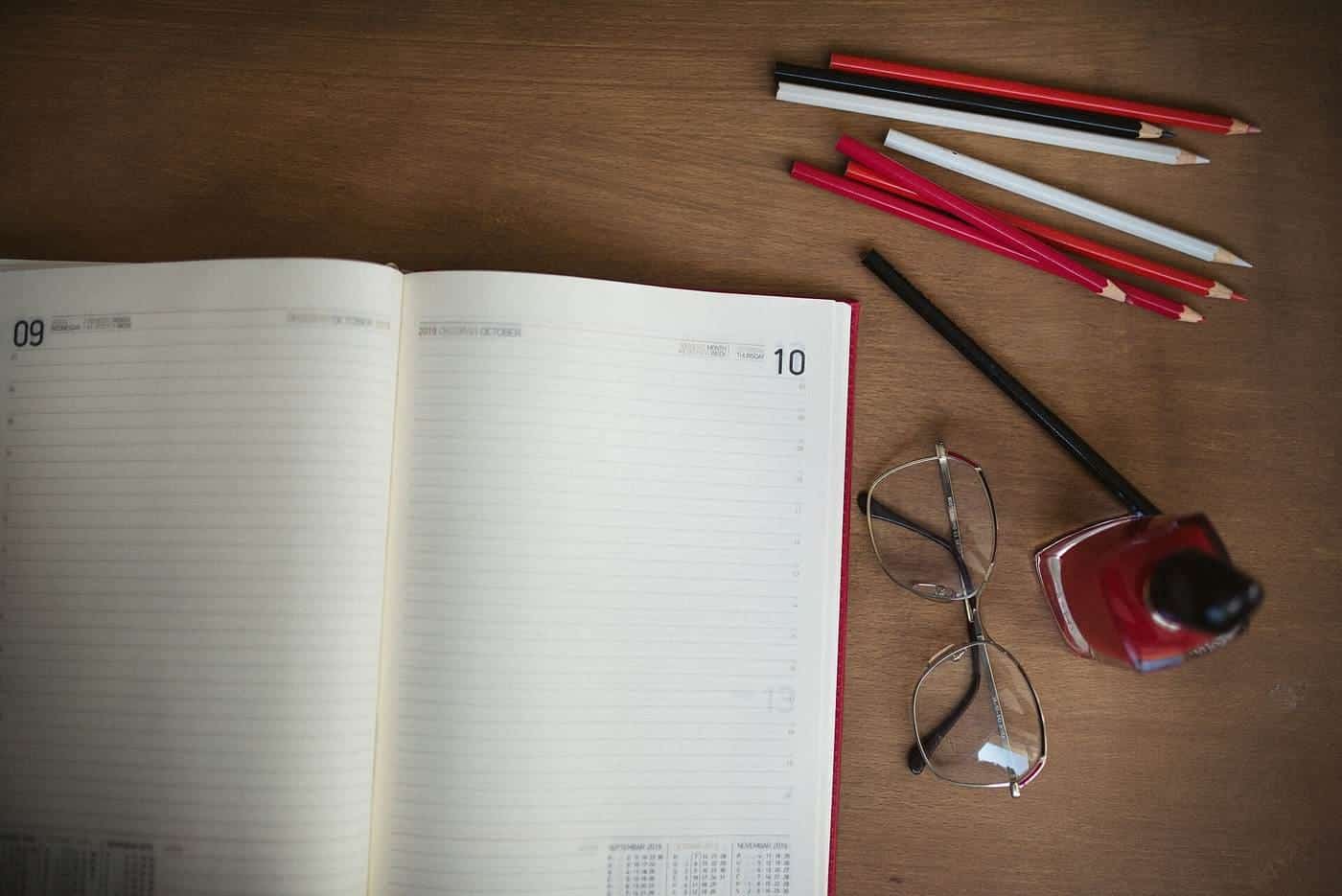 An open diary with pencils and reading glasses on a timber desk