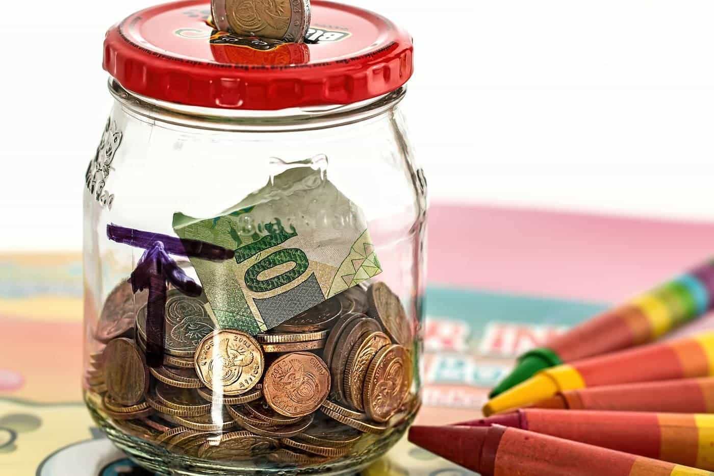 A glass jar of coins and notes beside colourful crayons