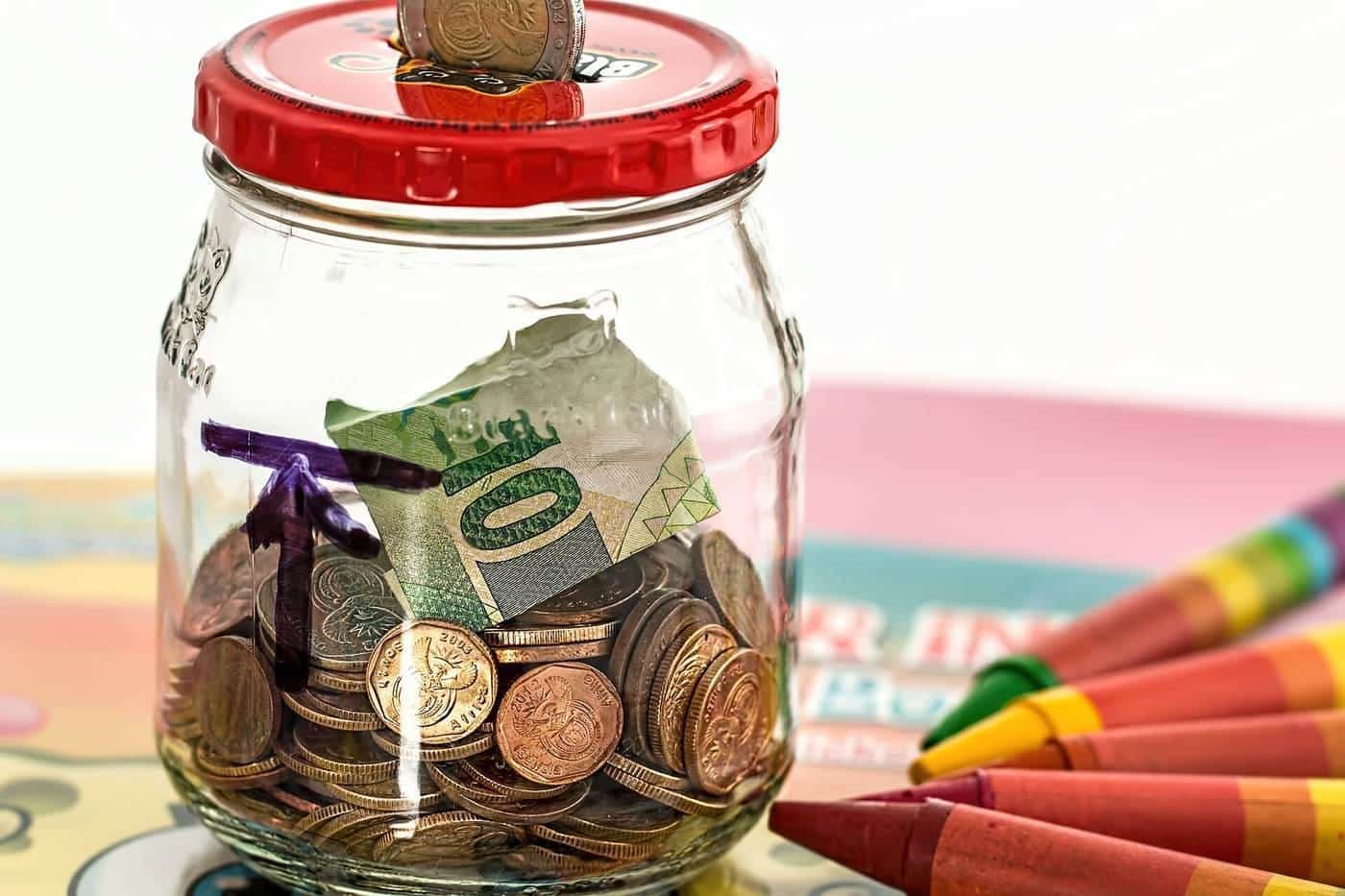 A glass jar of coins and notes beside colourful crayons